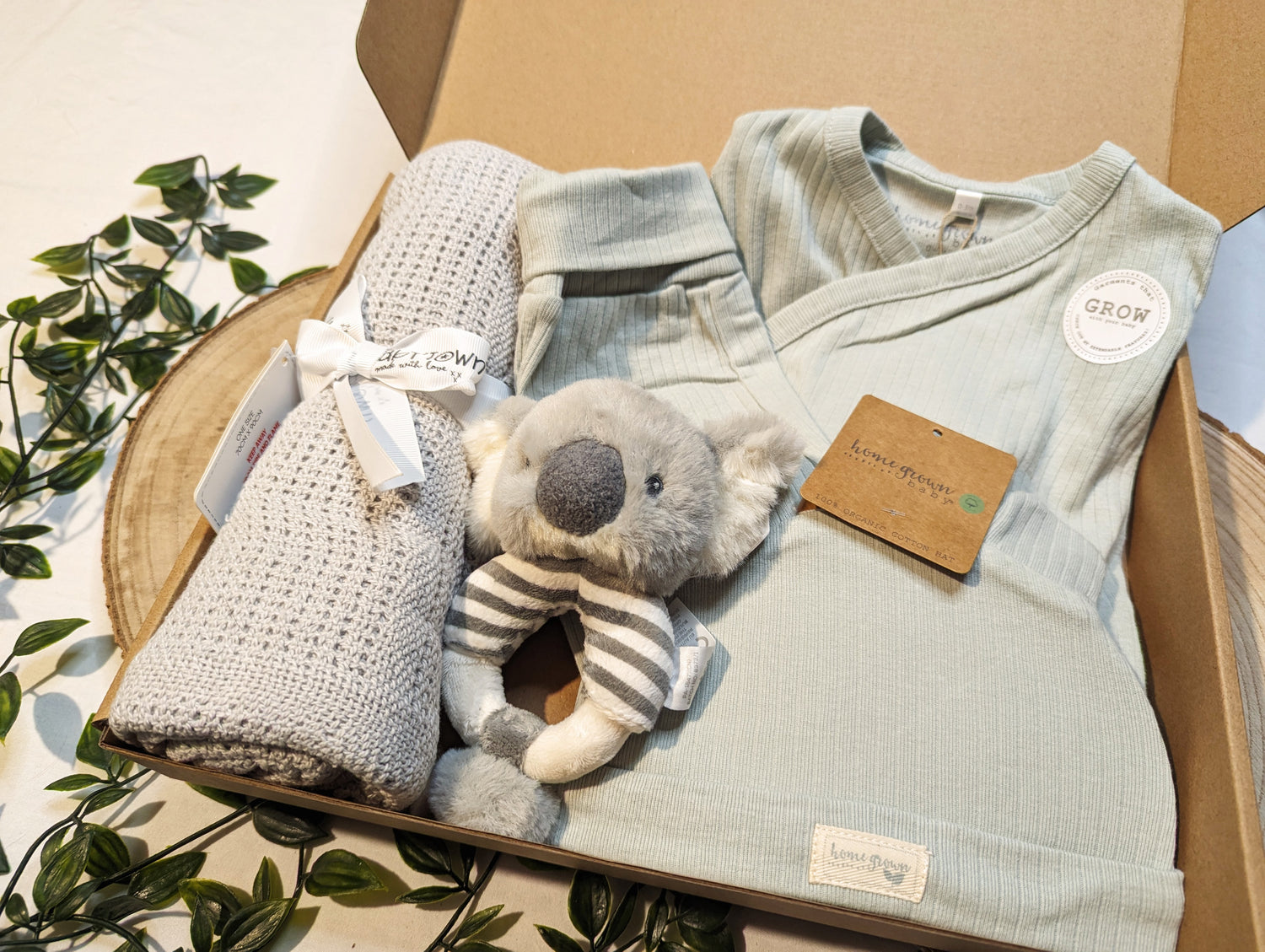 Unisex baby clothing gift packaged in a brown cardboard presentation box, includes a grey cellular baby blanket, baby koala soft ring rattle and three piece organic baby clothing outfit in sage green. The three piece baby clothing set includes one pair of ribbed trousers, one bodysuit and one hat with bear ears.  